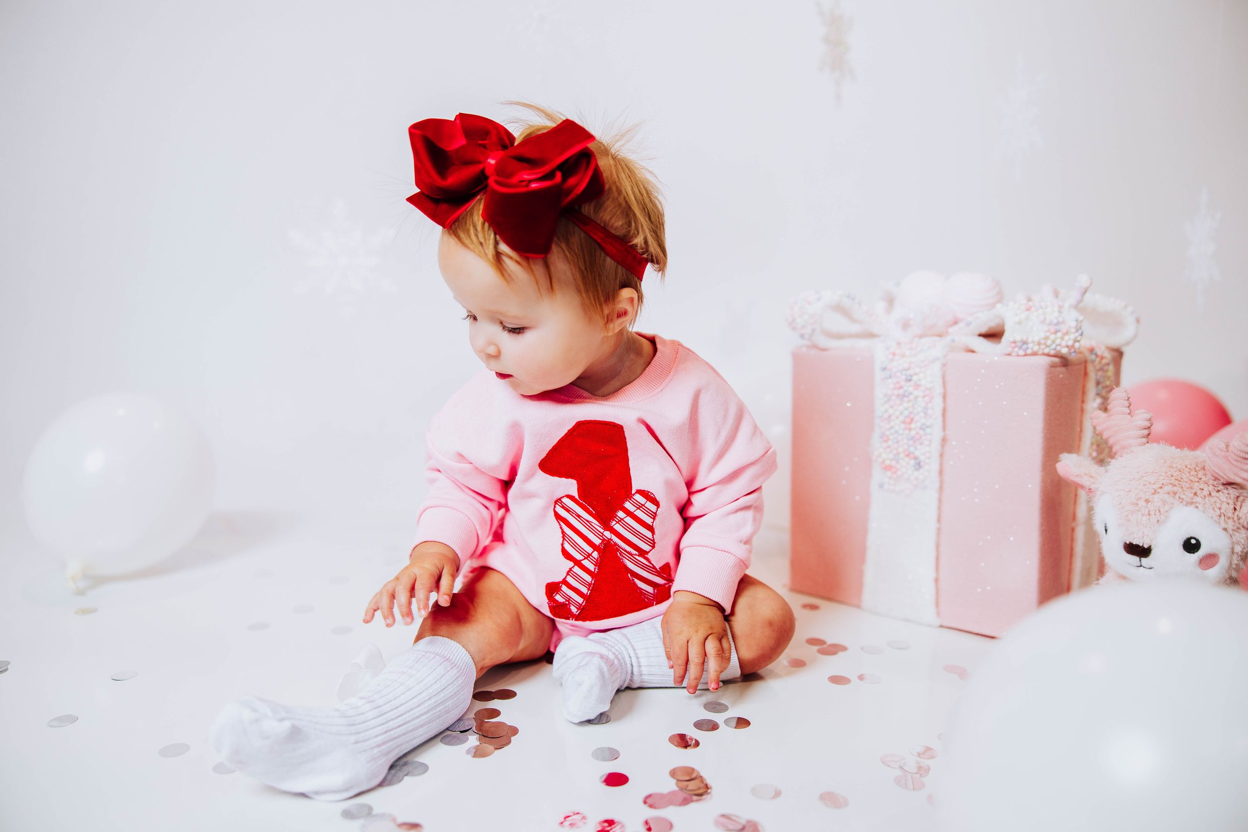 Baby girl in pink number one outfit with red bow headband sitting near gifts and balloons at first birthday.