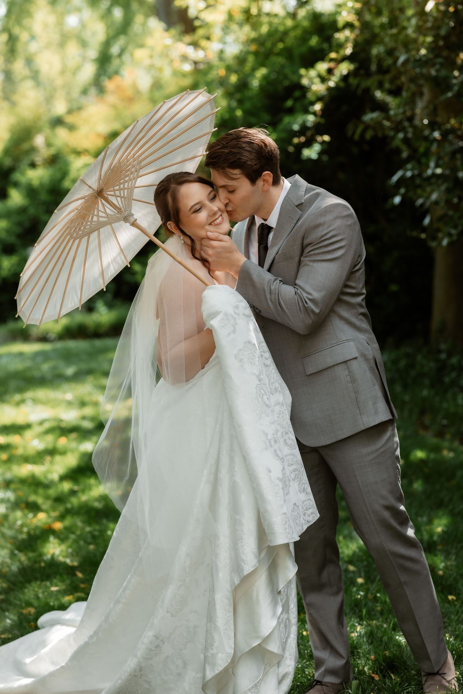 A romantic wedding couple shares an intimate moment under a white parasol in a lush garden setting.