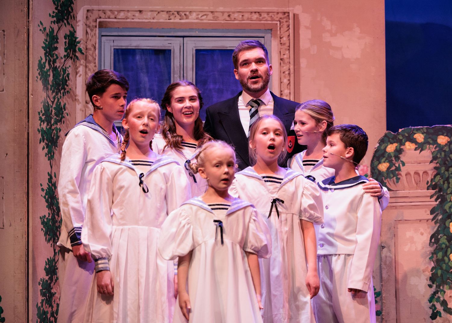 A group of performers in white choir robes stand together on stage during a theatrical production.