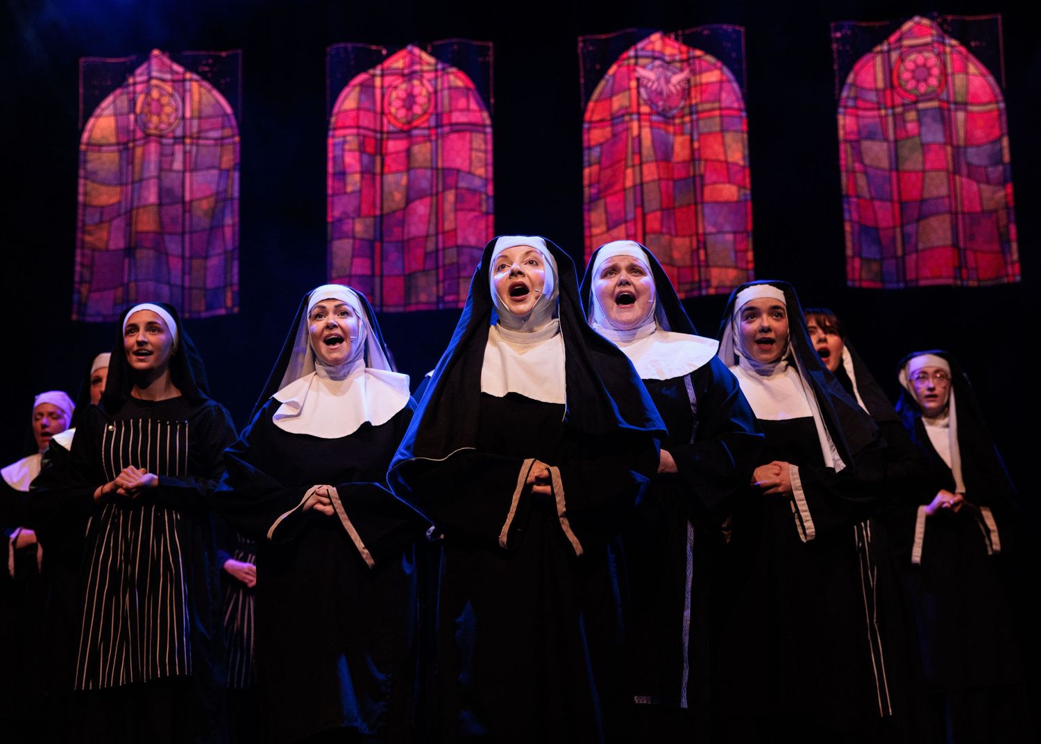Performers in black and white nun costumes stand on a dark stage illuminated by pink and purple theatrical lighting.