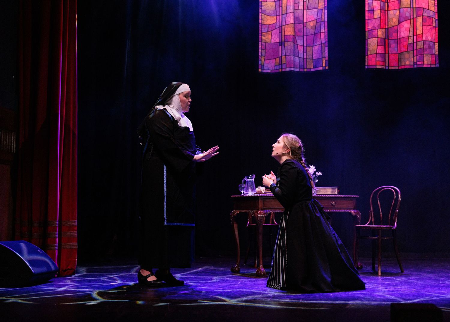 Two performers in black attire act out an emotional scene on a dimly lit theater stage with red lighting effects.