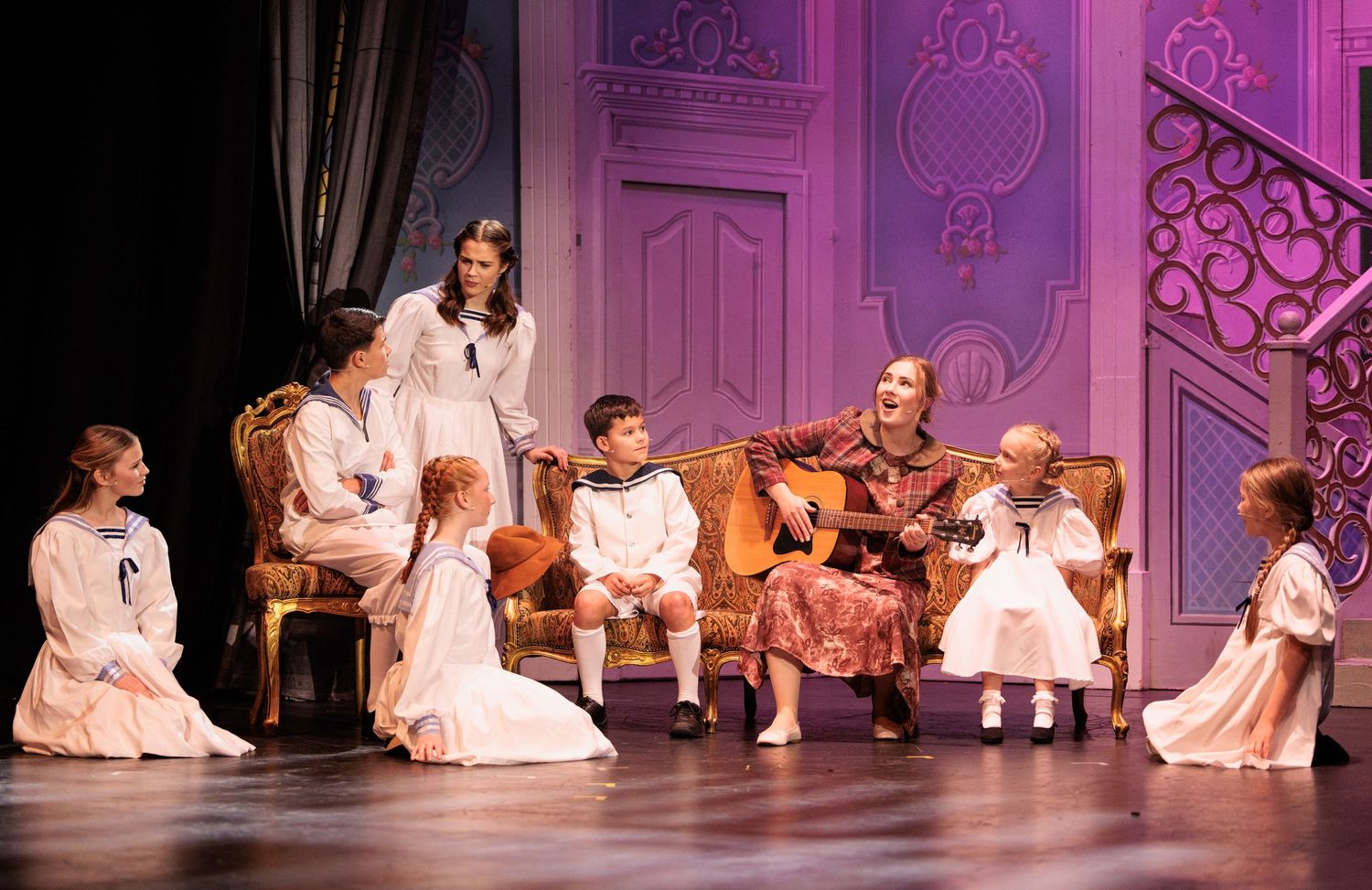 Performers in white dresses dance gracefully across a pink-lit stage during a theatrical production.