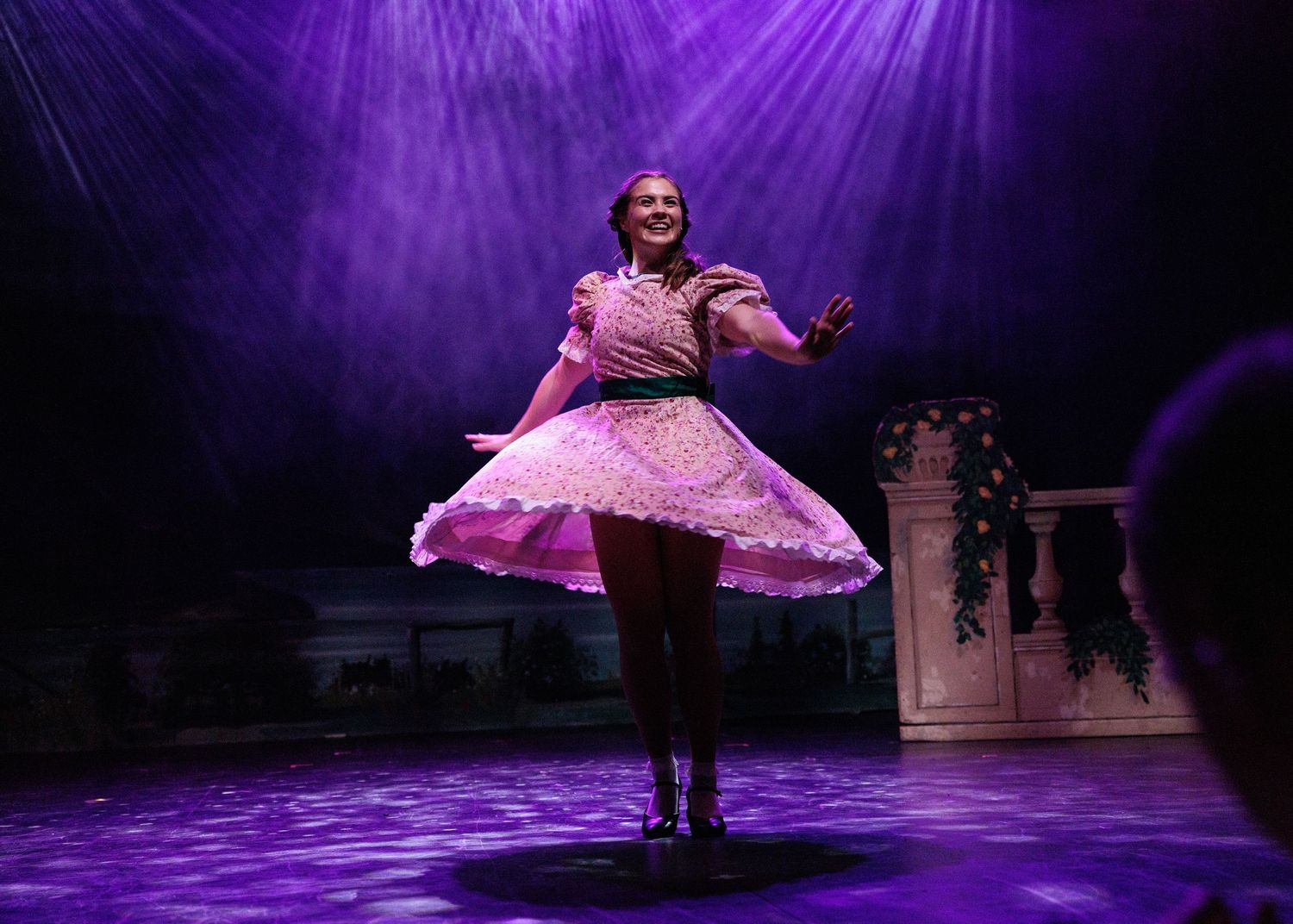A stage performance illuminated by purple lighting shows a dancer in motion during a theatrical production.