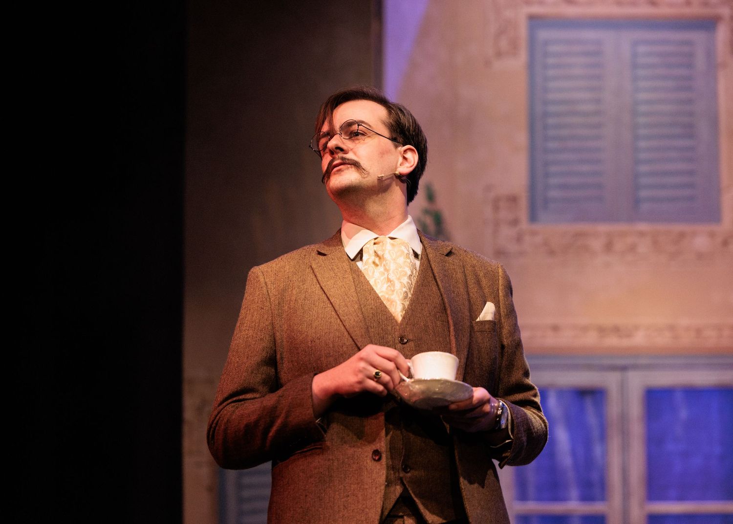 A performer in a brown suit gestures expressively while acting on stage with dramatic blue lighting in the background.