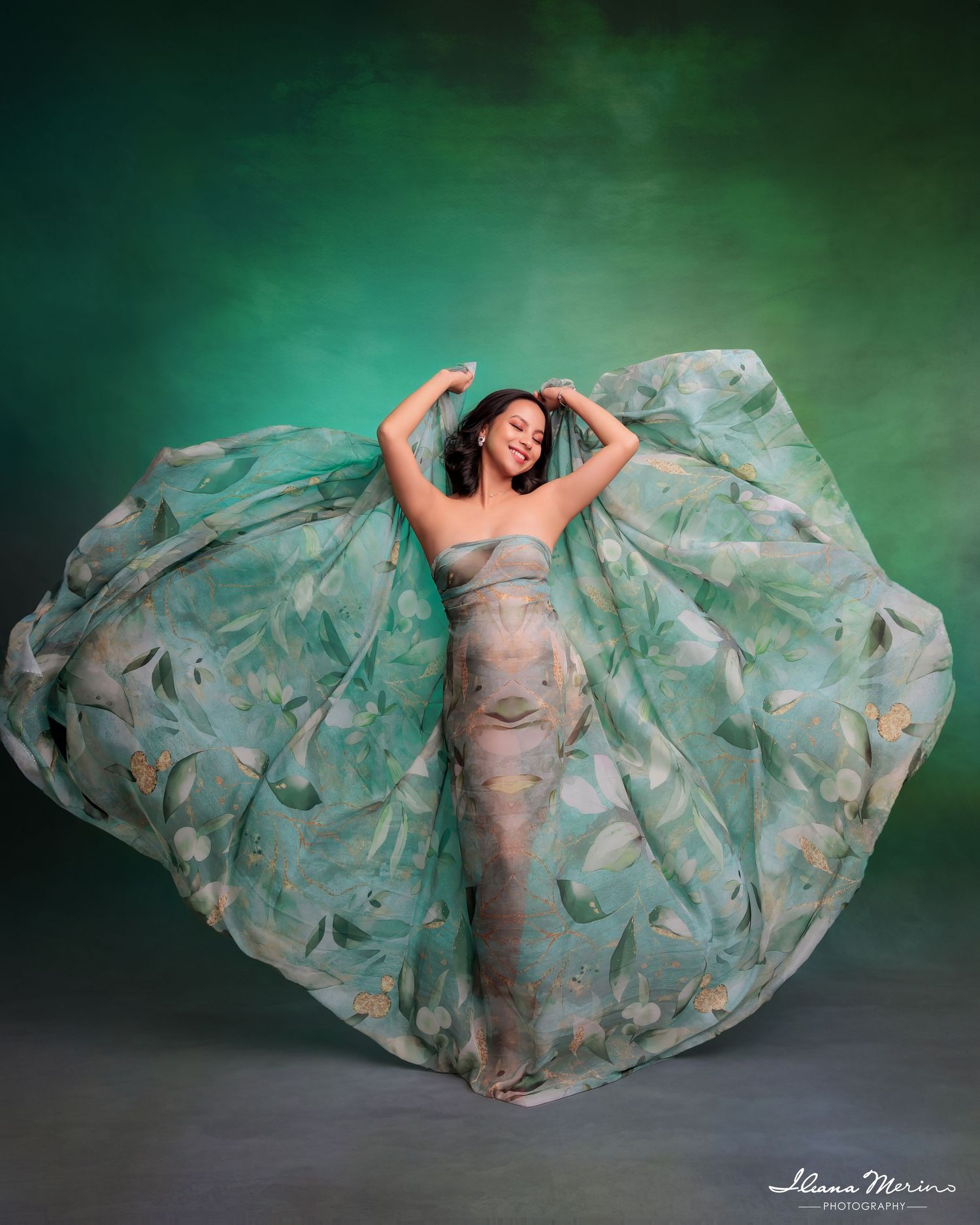 Pregnant woman twirls in flowing green floral chiffon gown against dramatic teal studio backdrop.