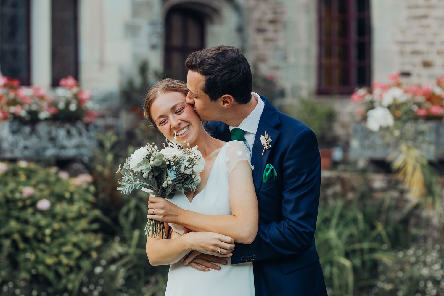 Smiling bride holding bouquet embraced by groom in navy suit outside flower-adorned stone building.