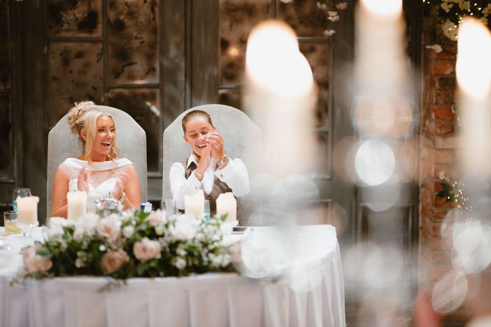 Two brides laughing joyfully at their wedding reception table adorned with candles and pink floral arrangements at Ballymagarvey Village