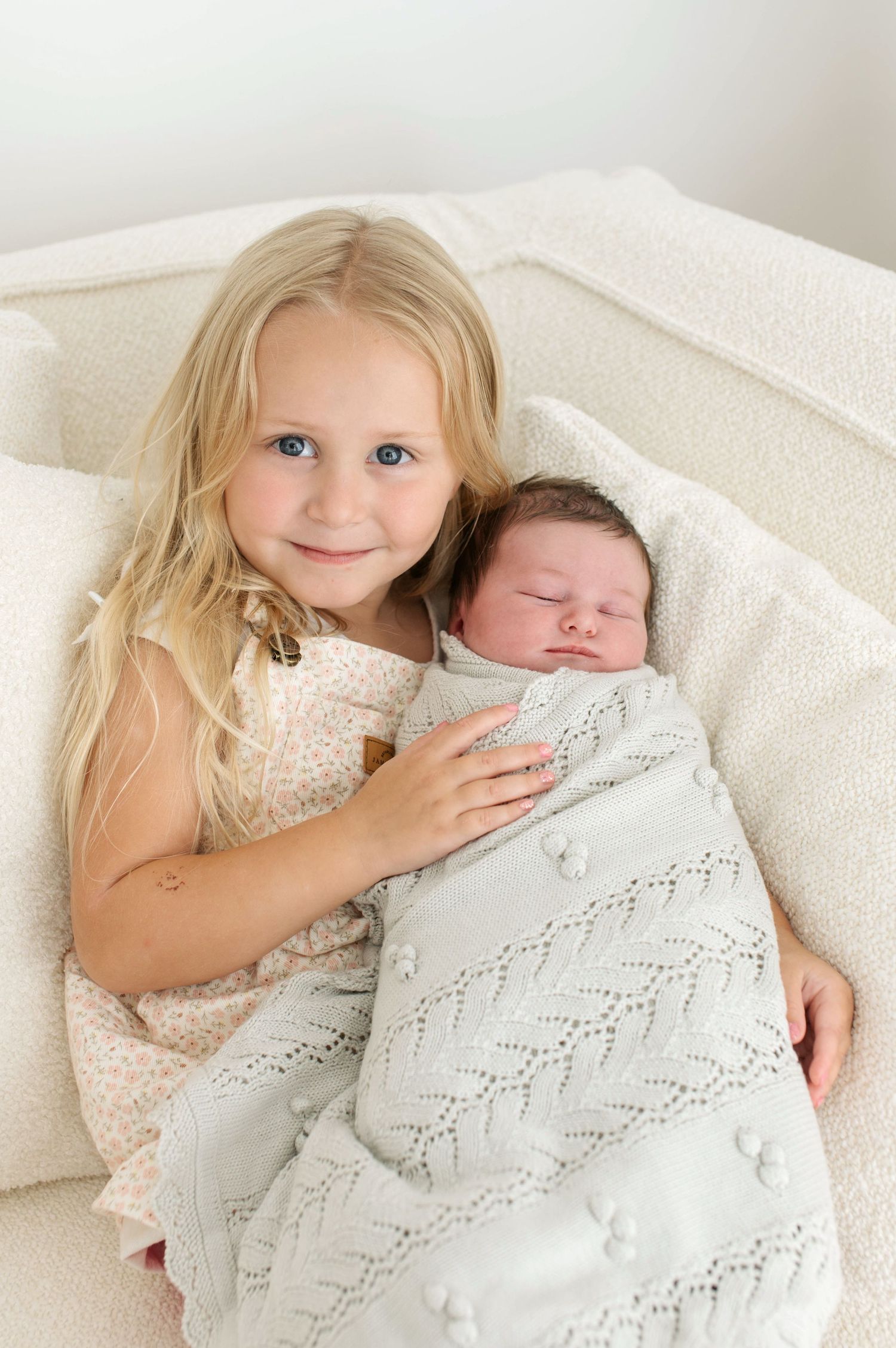A young child in a lace dress cuddles with a swaddled baby on white bedding in a series of sweet sibling photos.