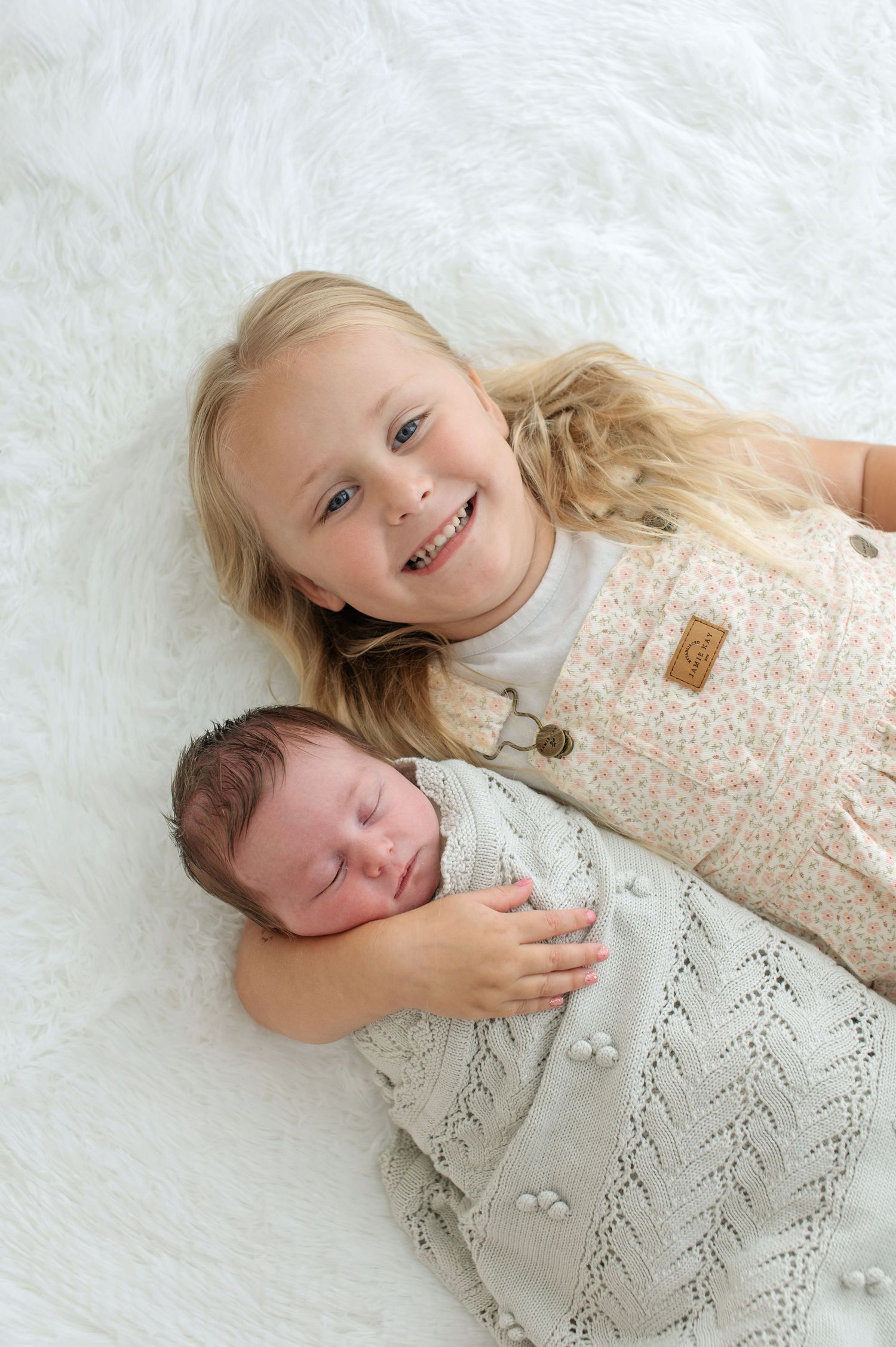Two children lying on white fur, with one wearing a cream knit sweater while the other sleeps in a gray knitted blanket.