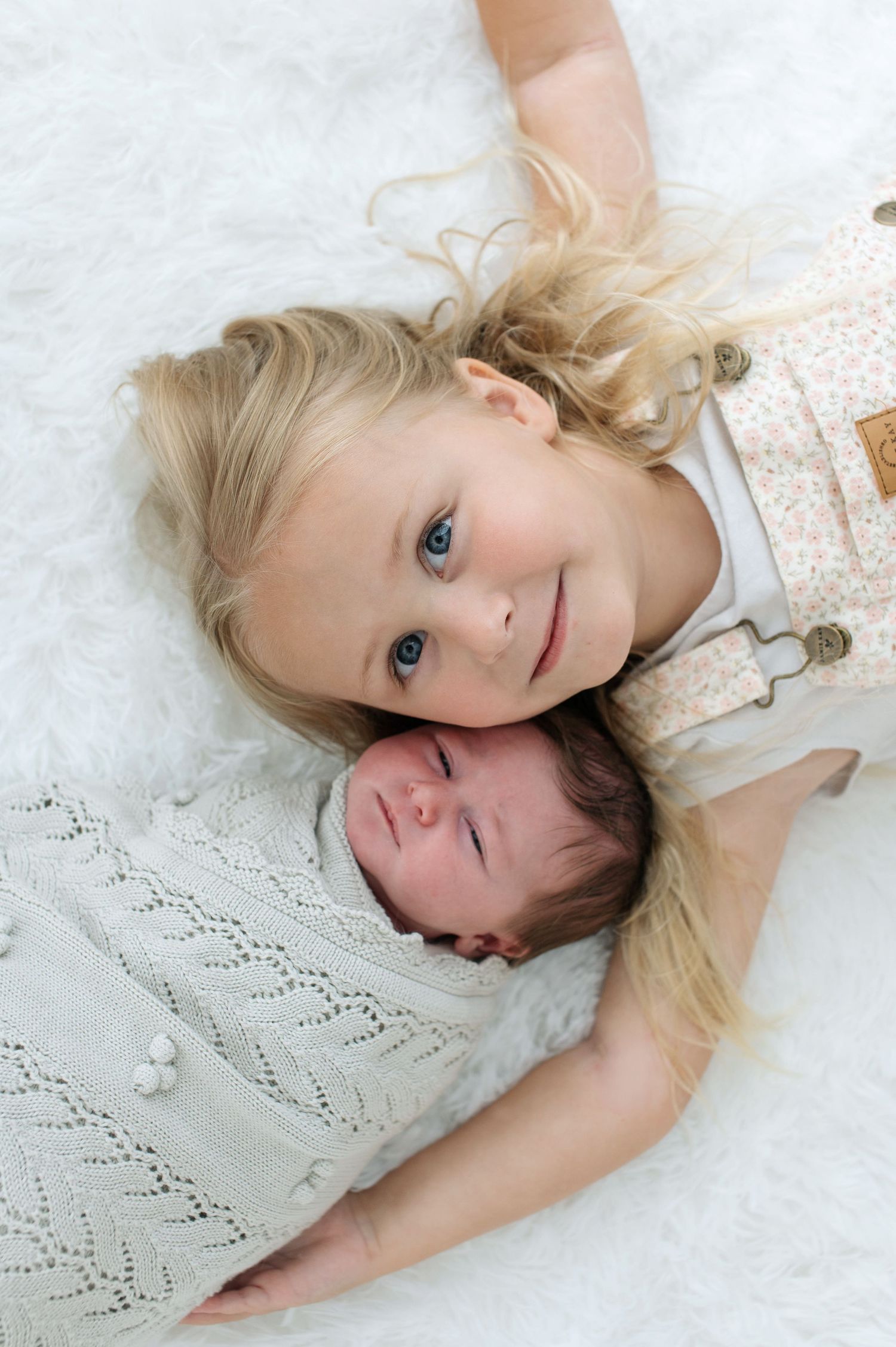 Two children laying on white bedding with knit blanket, one older with blonde hair and one infant.