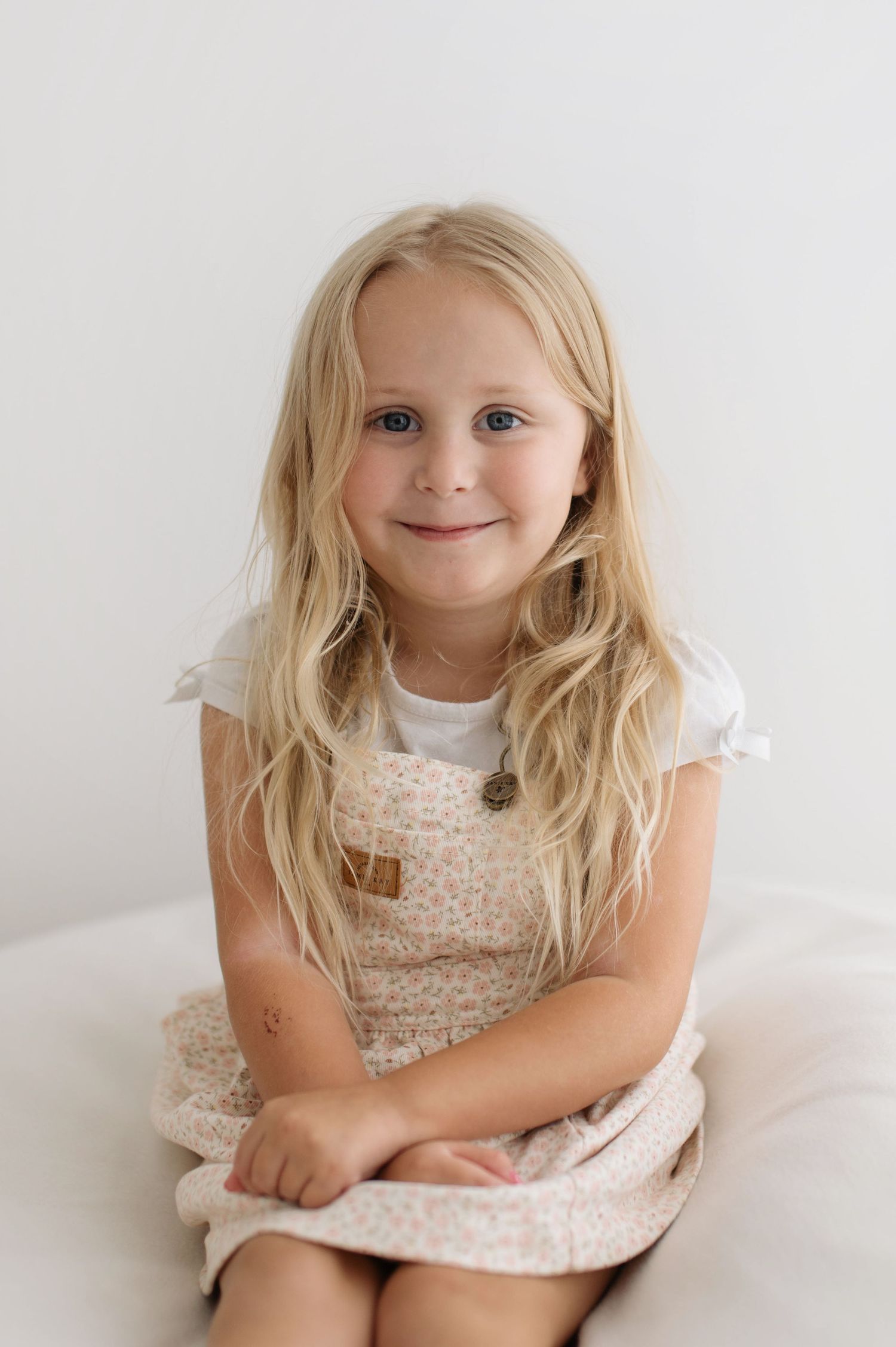 A young child in a white floral dress poses against a light background with a sweet smile.