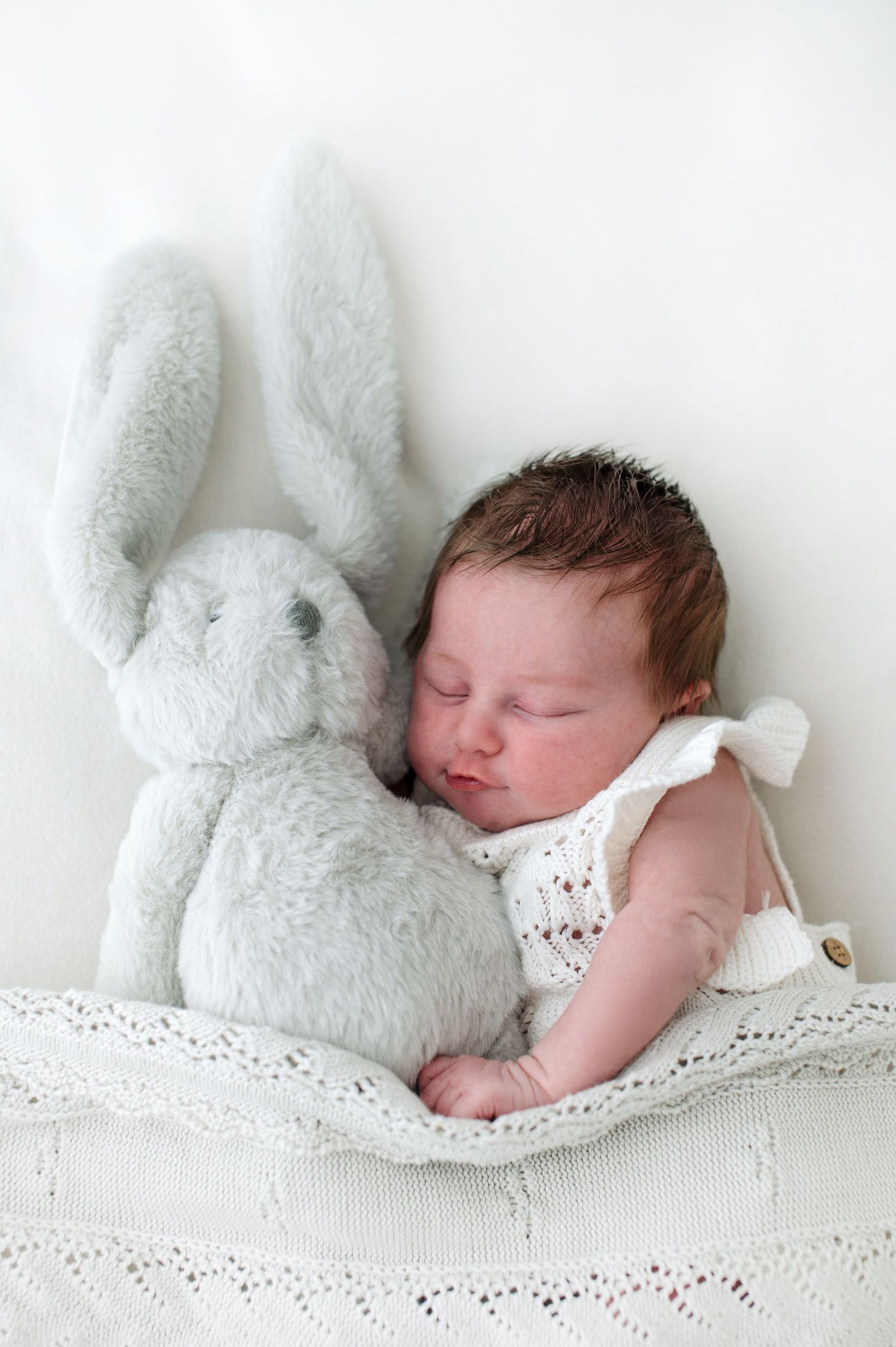 A sleeping newborn baby rests peacefully on white lace bedding next to a soft plush bunny toy.