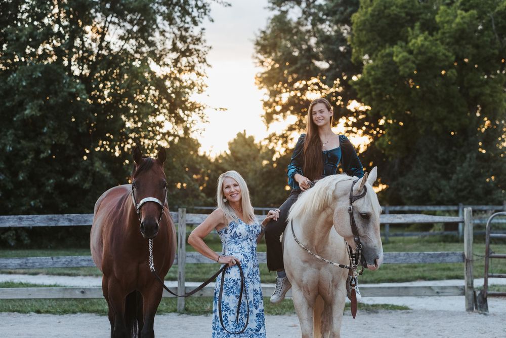 People and horses gather in a fenced paddock area during golden hour sunset lighting.
