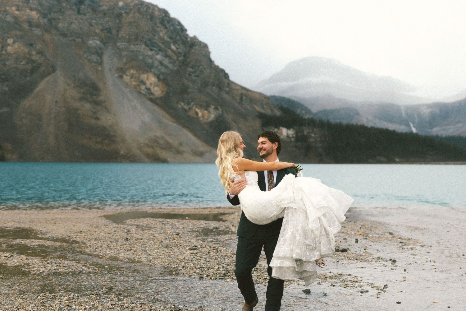 groom carries bride across a river in Banff and grins at her