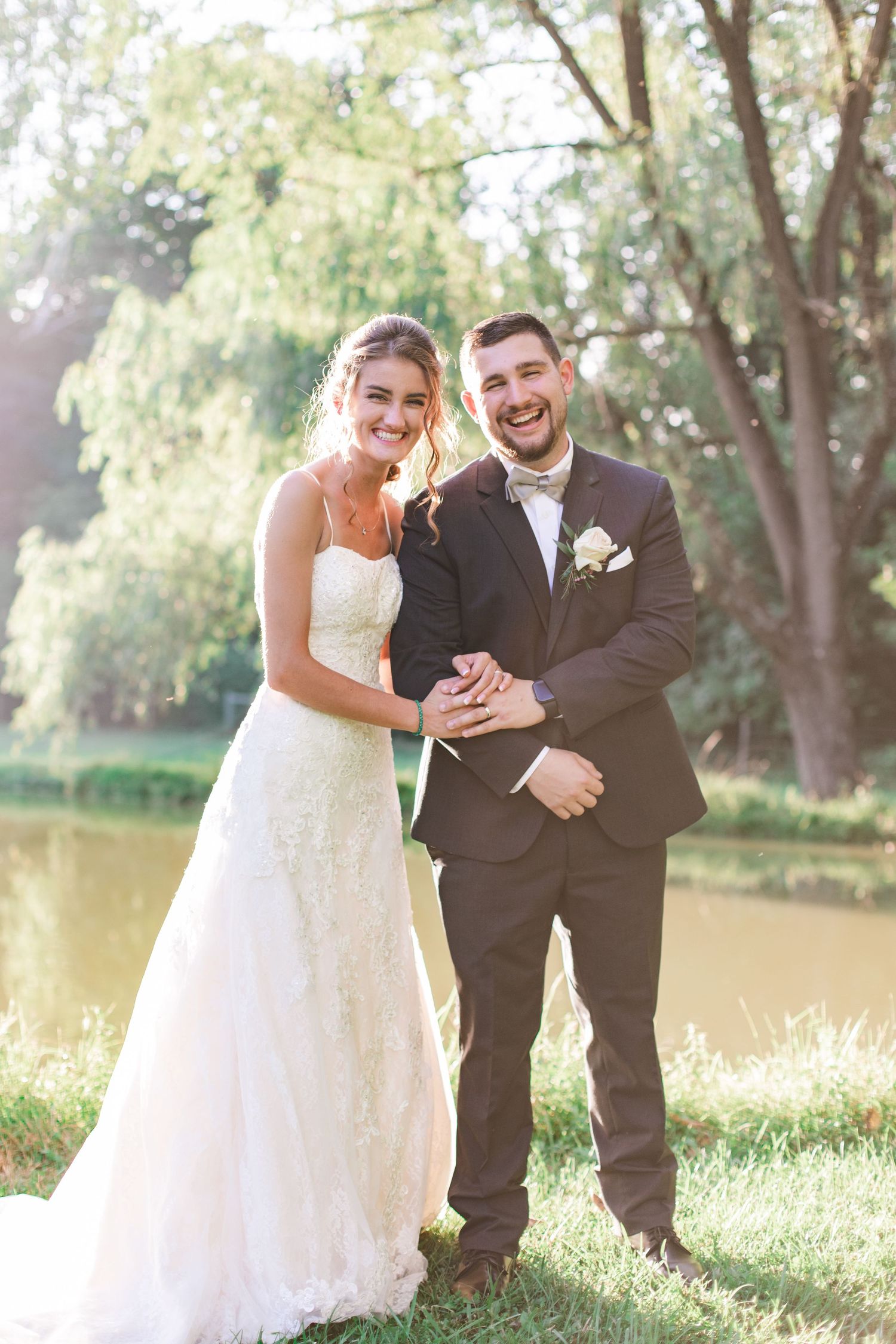 Laughing bride and groom pose together near a serene pond surrounded by weeping willow trees on a sunny day.