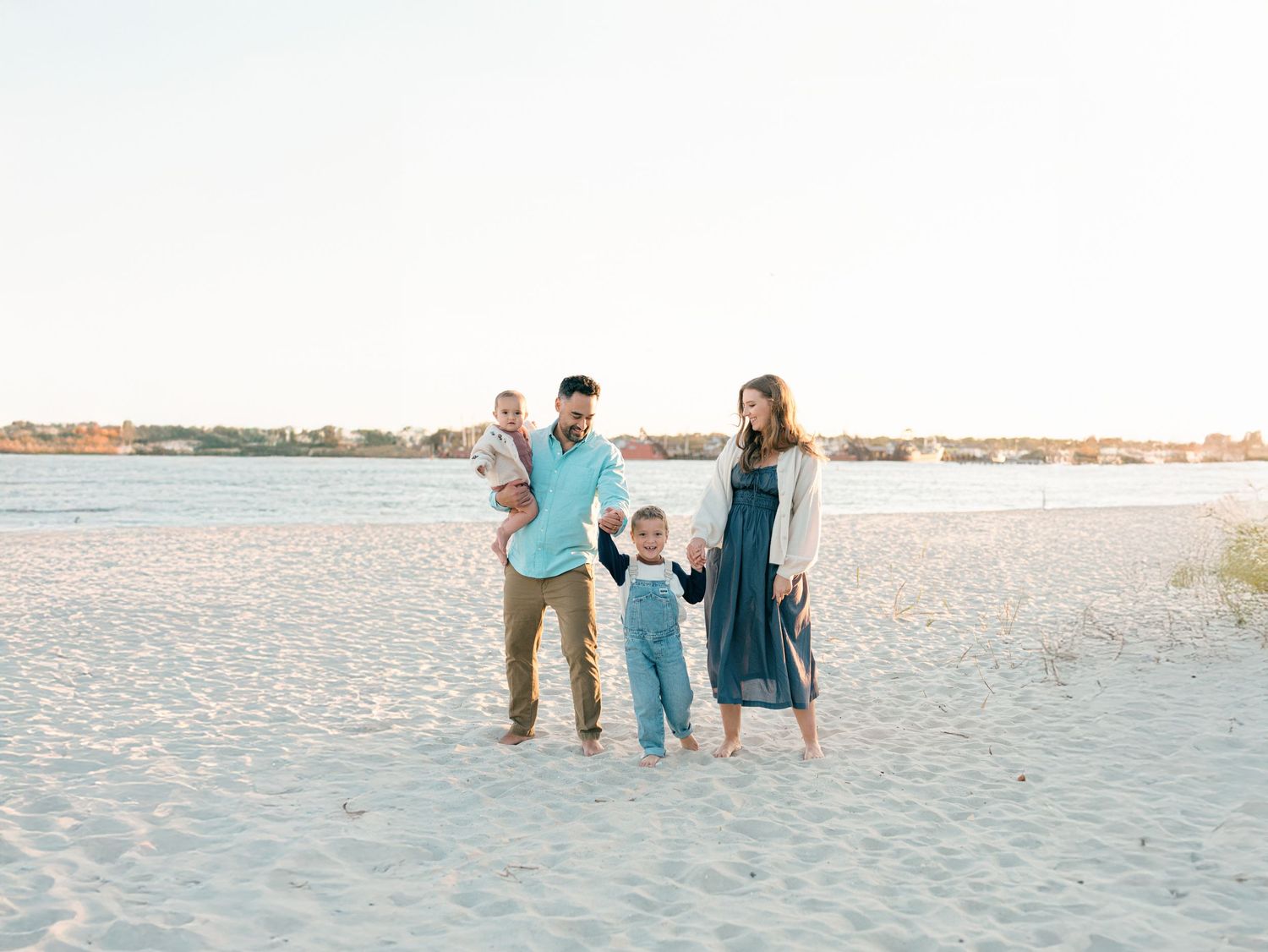 A family of four walks down the nj beach holding hands and smiling in blue and tan outfits.