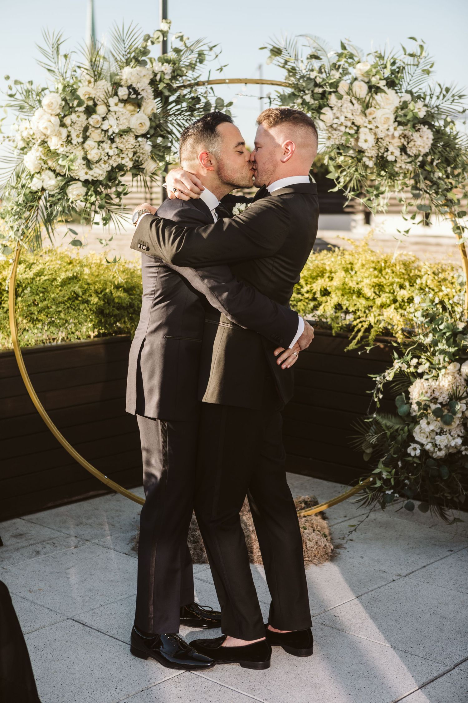 Two people in formal black attire embrace during an outdoor wedding ceremony with white floral decorations.
