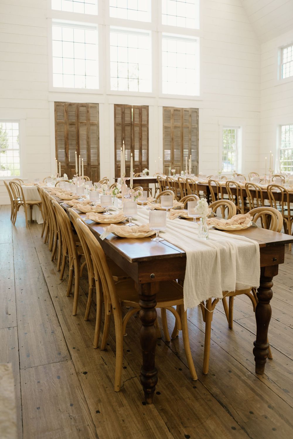 Long wooden farm tables set for a wedding reception in an elegant white room with natural light.