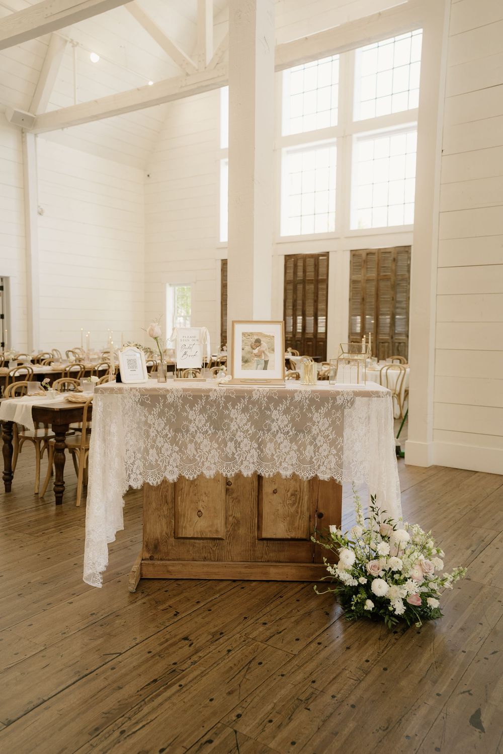 White reception hall with wooden floors and lace-draped tables decorated for a wedding.