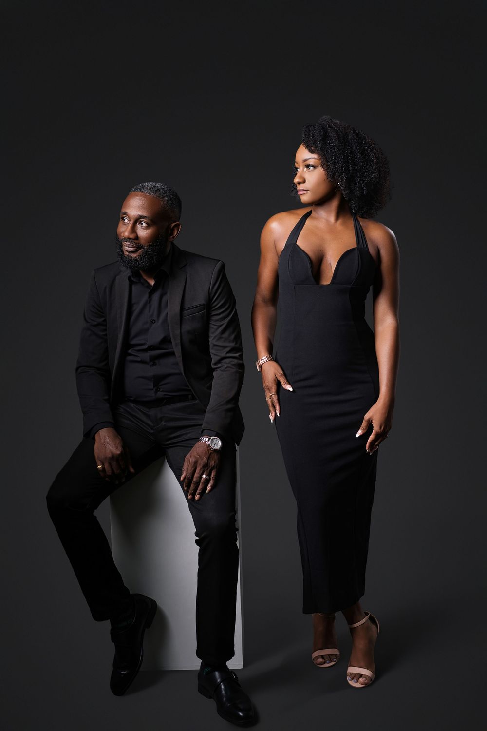 A couple in all black outfits poses together against a dark background in a dramatic studio photoshoot.