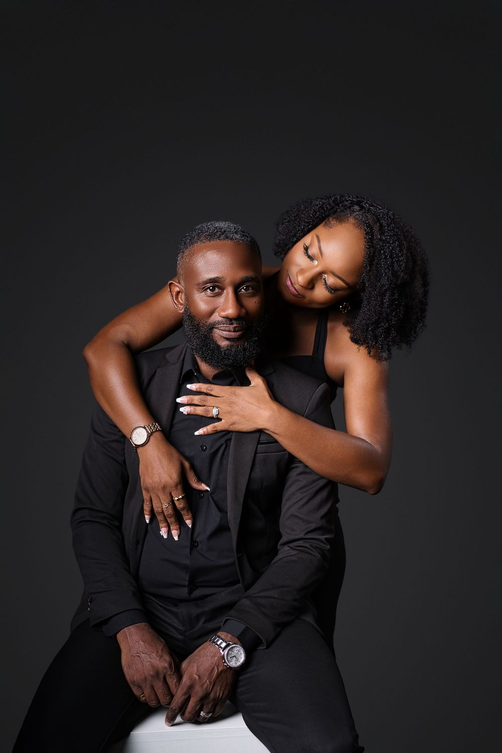 Intimate couple portrait in black attire against dark backdrop showing tender embrace and emotional connection.