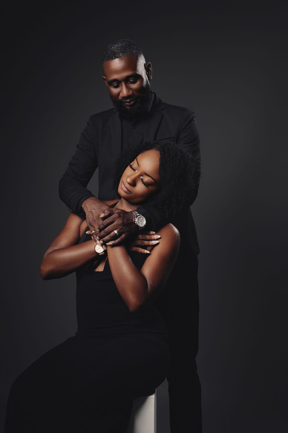 Romantic studio portrait showing intimate embrace between couple in black against dramatic dark background.