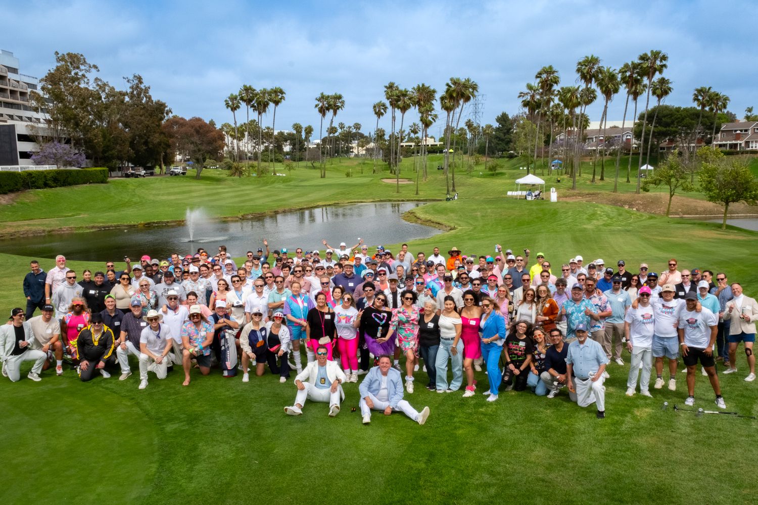 Large group of golf tournament participants gathered on a lush green course with palm trees in the background.