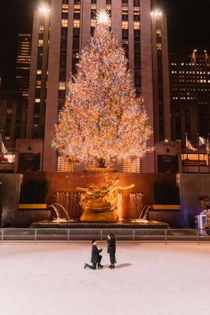 Marriage proposal in front of illuminated Christmas tree at Rockefeller Center ice rink.