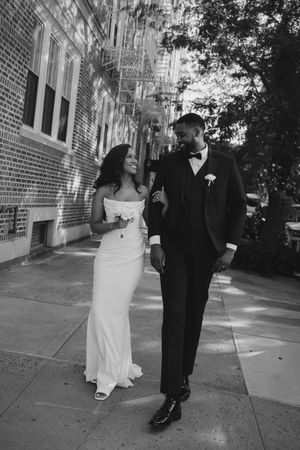 A couple in wedding attire walks together on a brick-lined sidewalk in black and white.