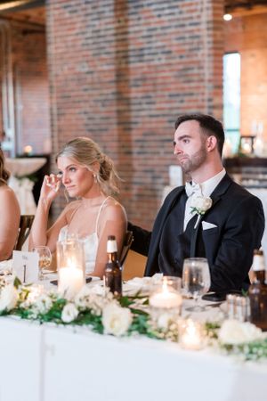 Wedding reception guests seated at an elegant long table with candlelight and greenery in a brick-walled venue.
