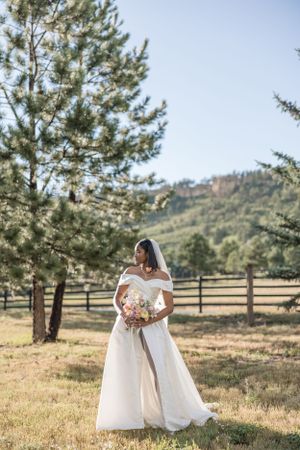 Bride in off-shoulder white gown holds bouquet in rustic mountain setting.
