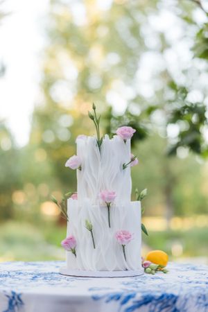 Elegant white wedding cake decorated with fresh pink flowers on blue patterned tablecloth.
