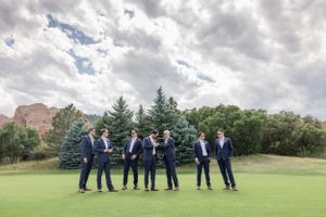 Groomsmen in navy suits pose for photos on a grassy hill against cloudy skies.