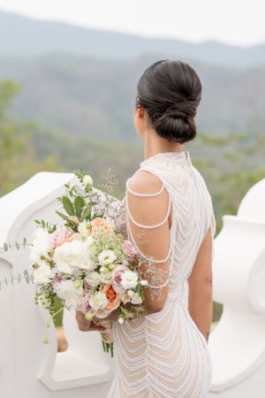Back view of a bridal gown with intricate beaded detail and floral bouquet against mountain backdrop.