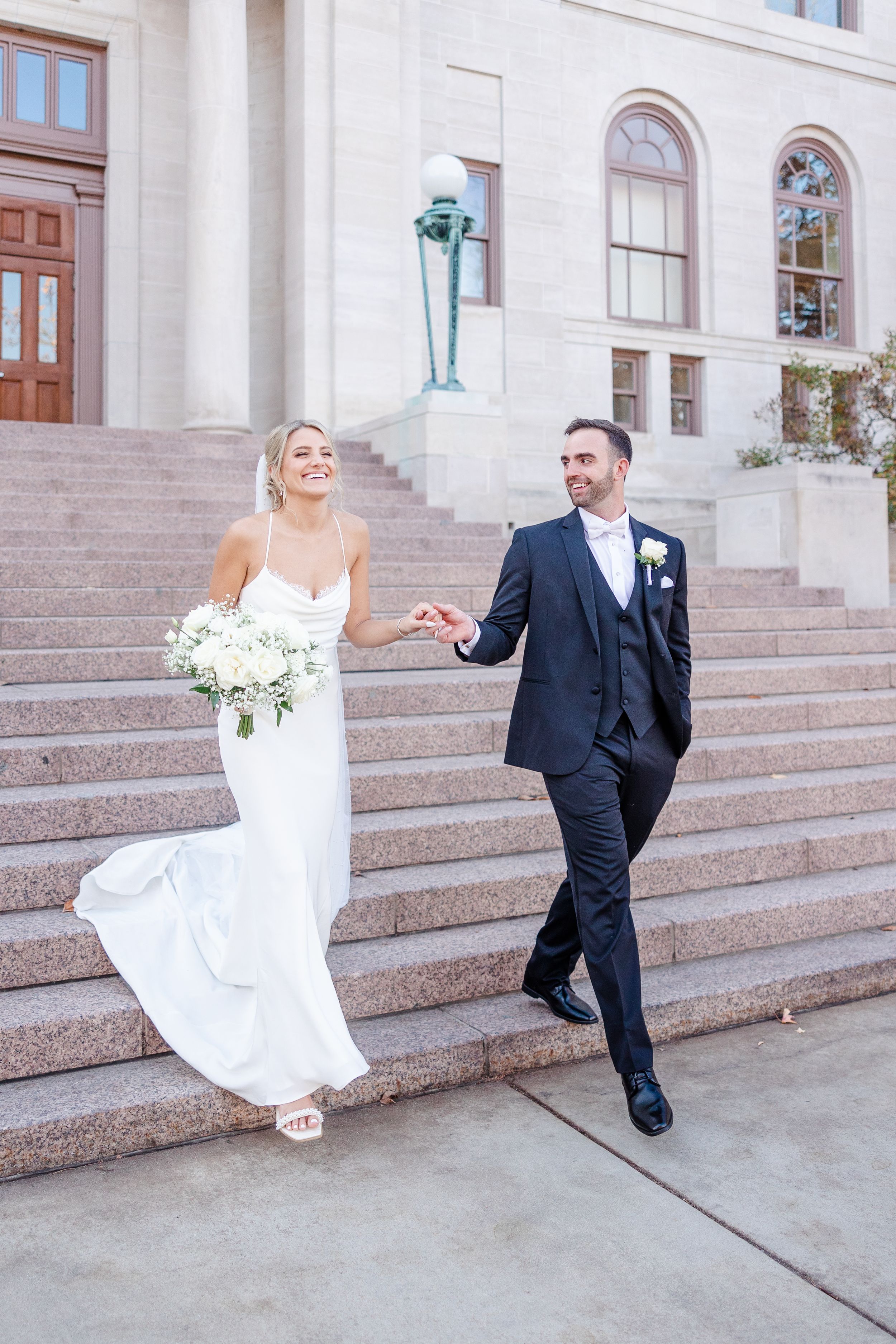 A couple descends stone steps outside a grand building in formal wedding attire.