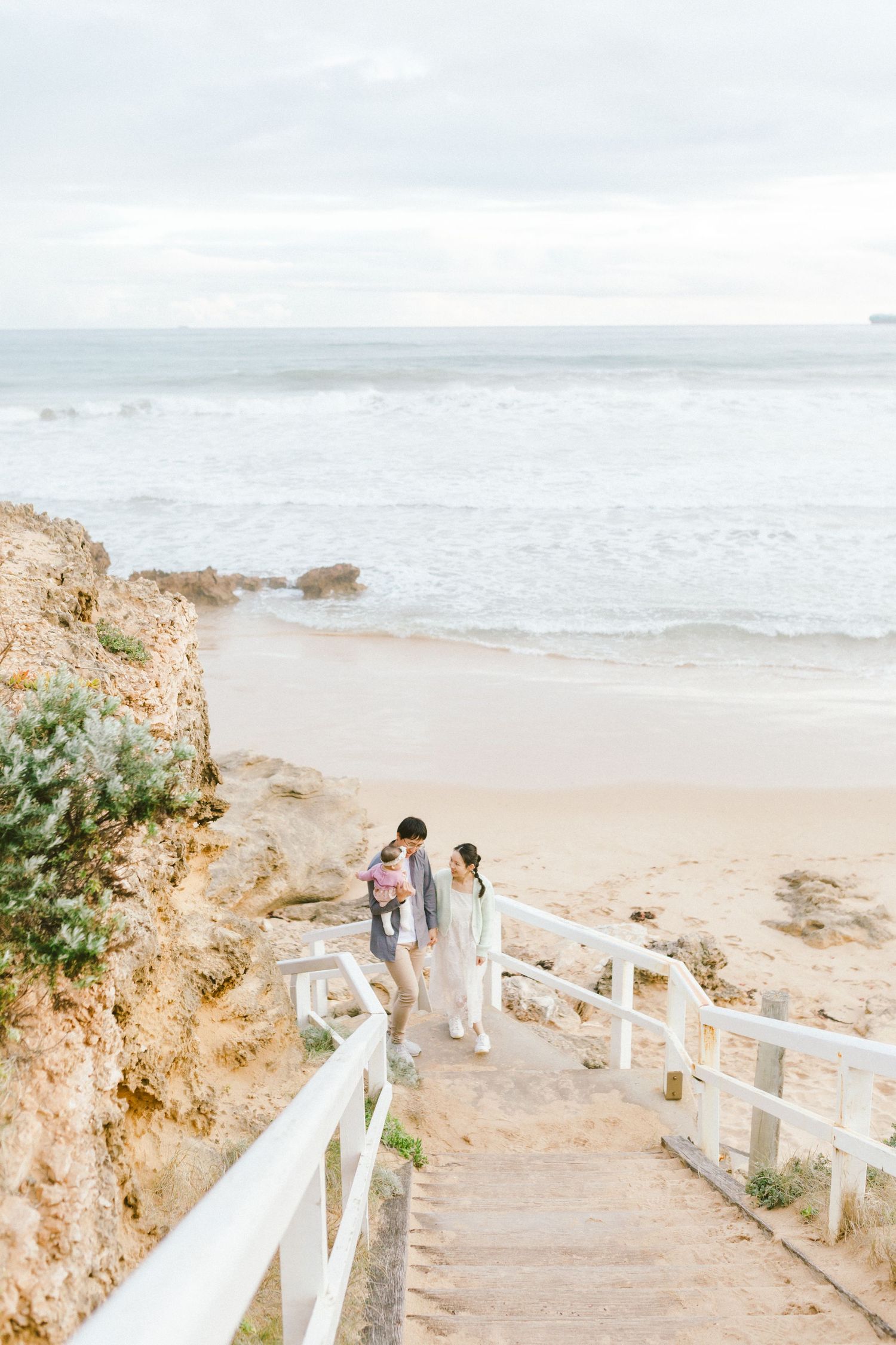 A couple walks down white wooden stairs leading to a scenic beach with cliffs and ocean waves in soft pastel tones.