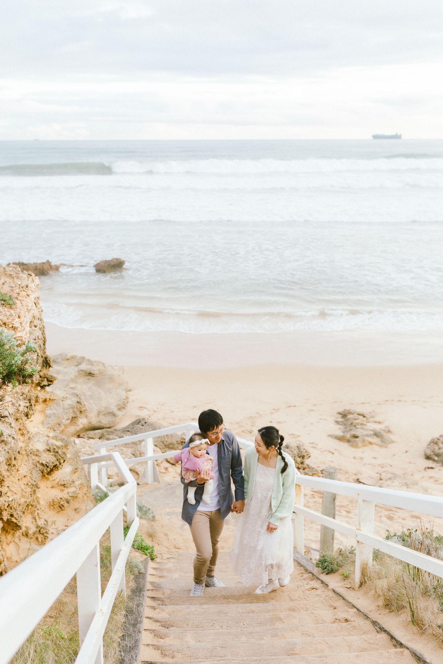 A couple walks down wooden stairs leading to a scenic beach with ocean views on an overcast day.