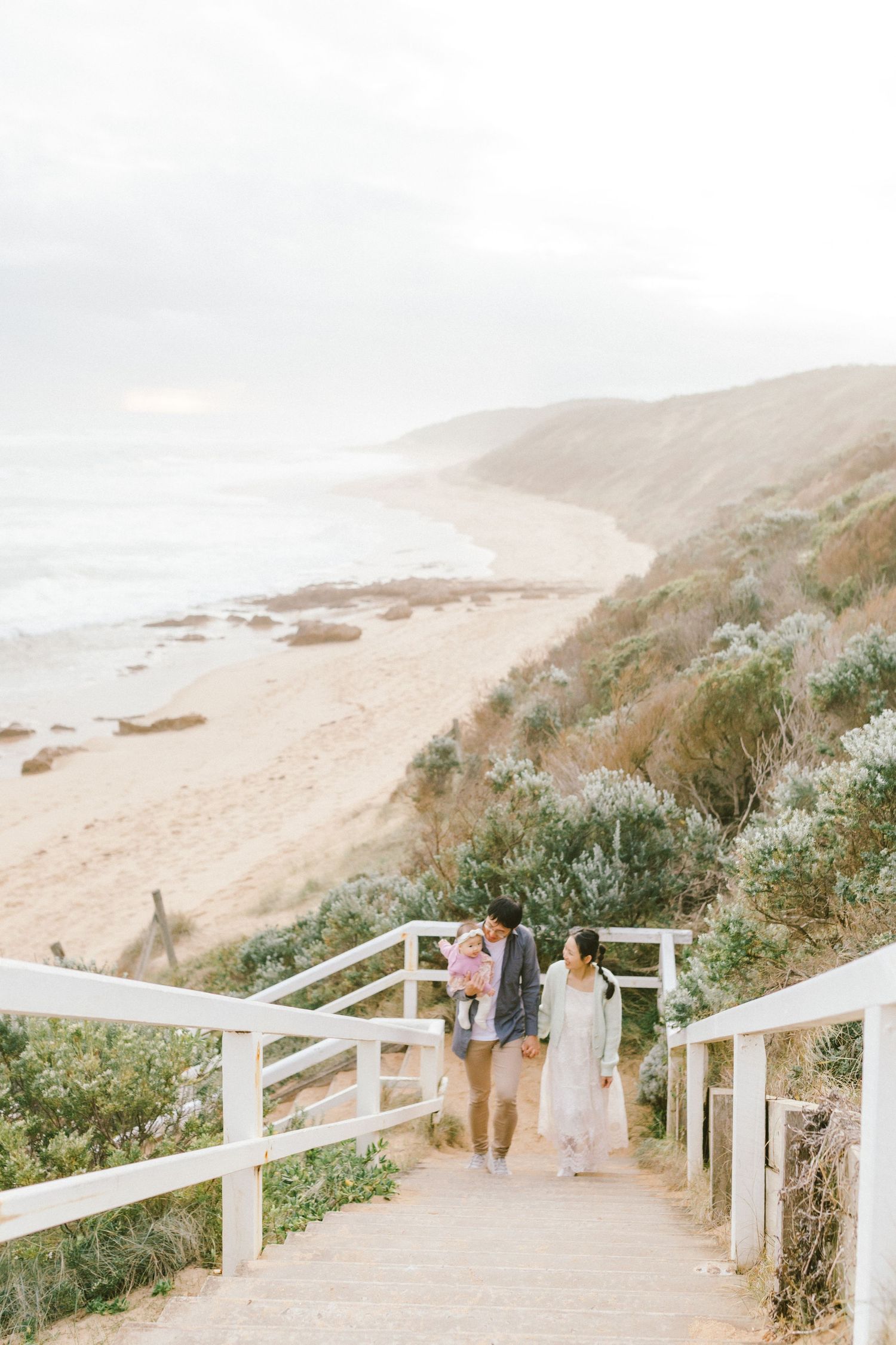 Couple walking down coastal boardwalk stairs overlooking scenic beach and cliffs on foggy day.