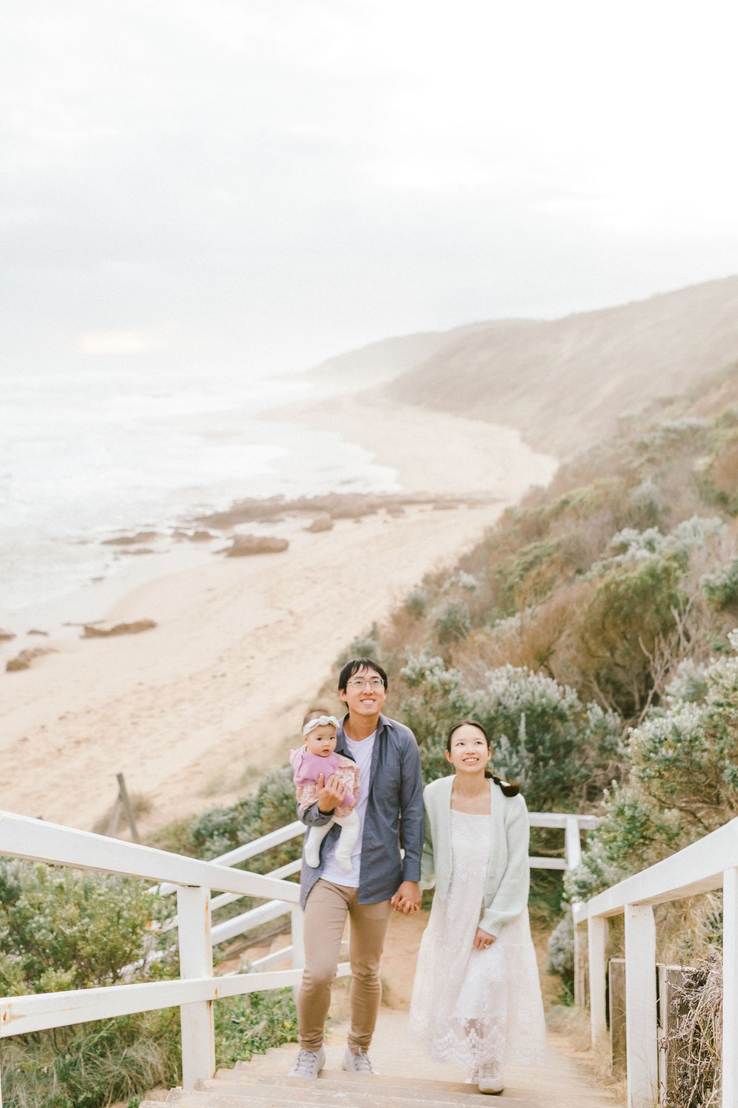 A couple walks along a scenic coastal path with white fencing and rolling hills in the background.