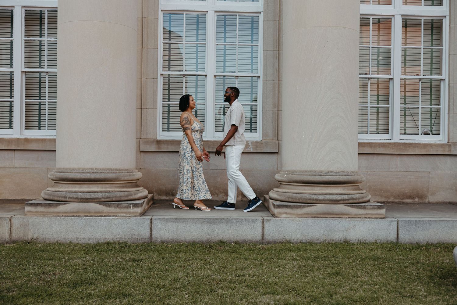 A couple in formal attire walks together on the lawn outside an elegant building with white columns and windows.