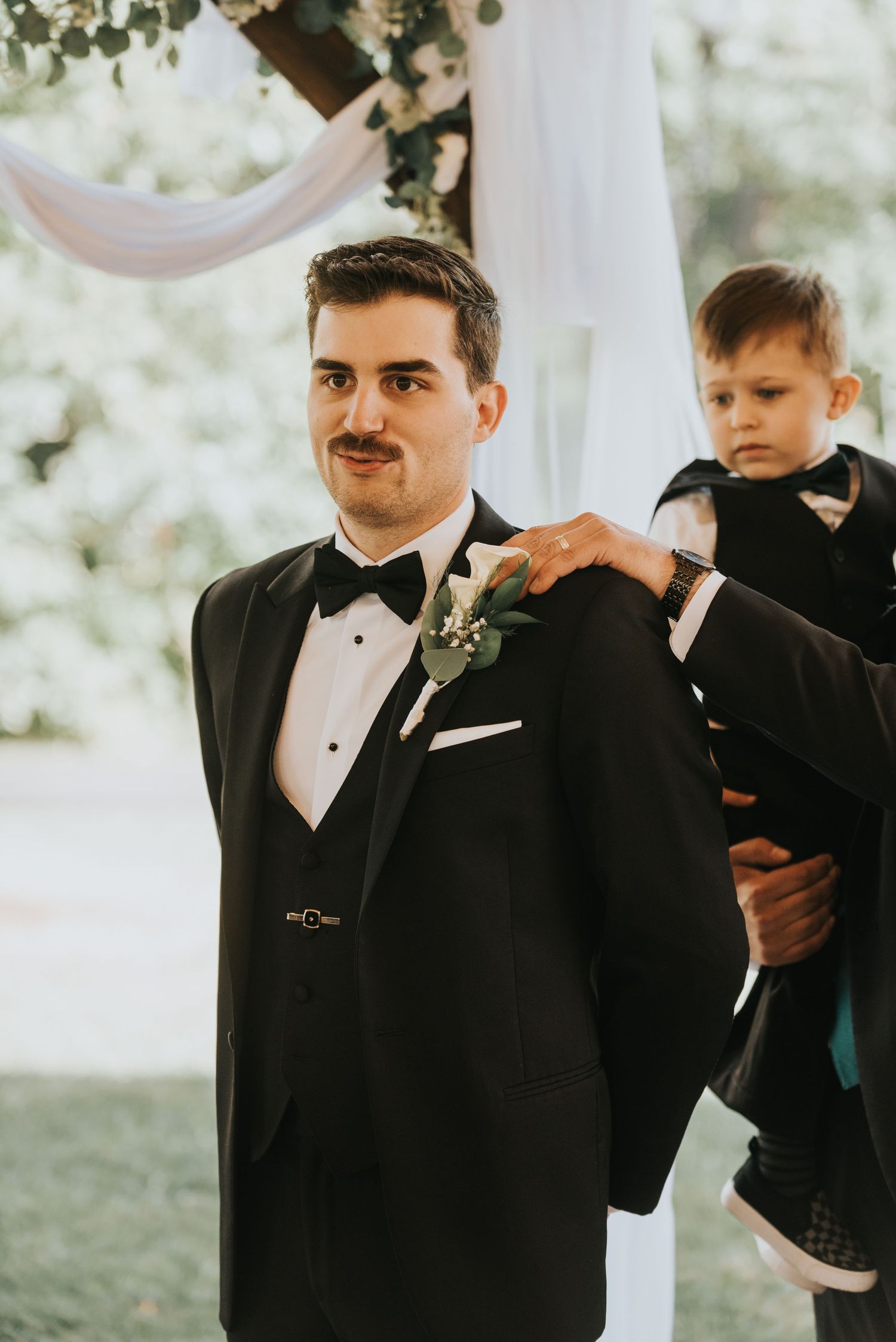 A groom in a black tuxedo and bowtie adjusts his tie while standing outdoors at a wedding ceremony.