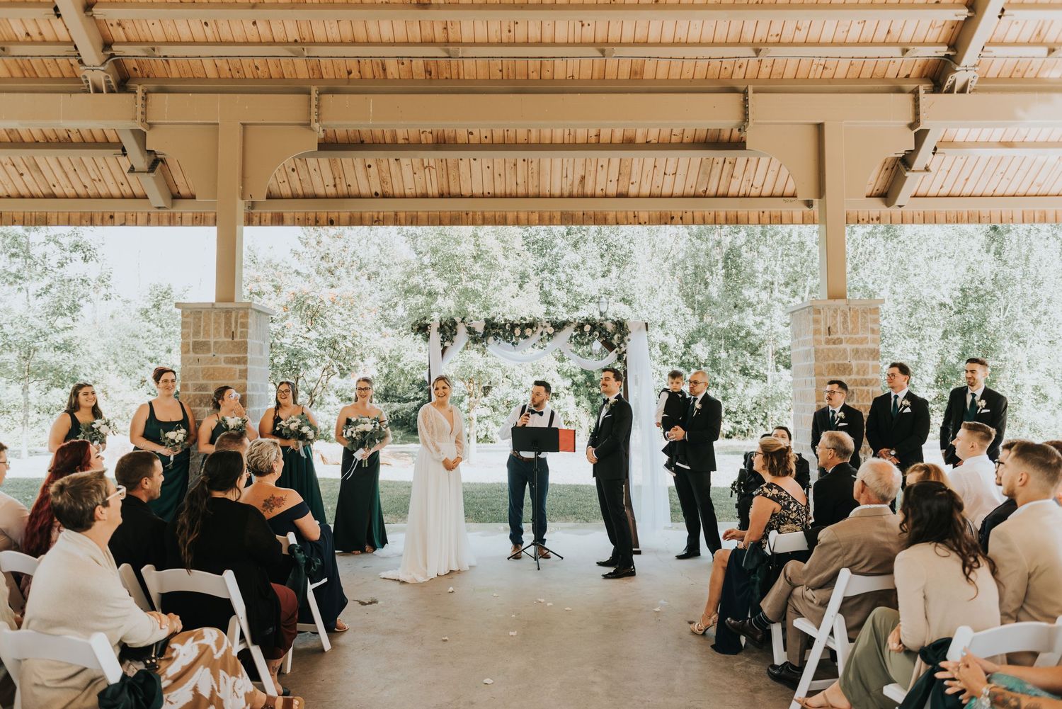 Wedding guests seated in rows at an outdoor covered pavilion ceremony space.
