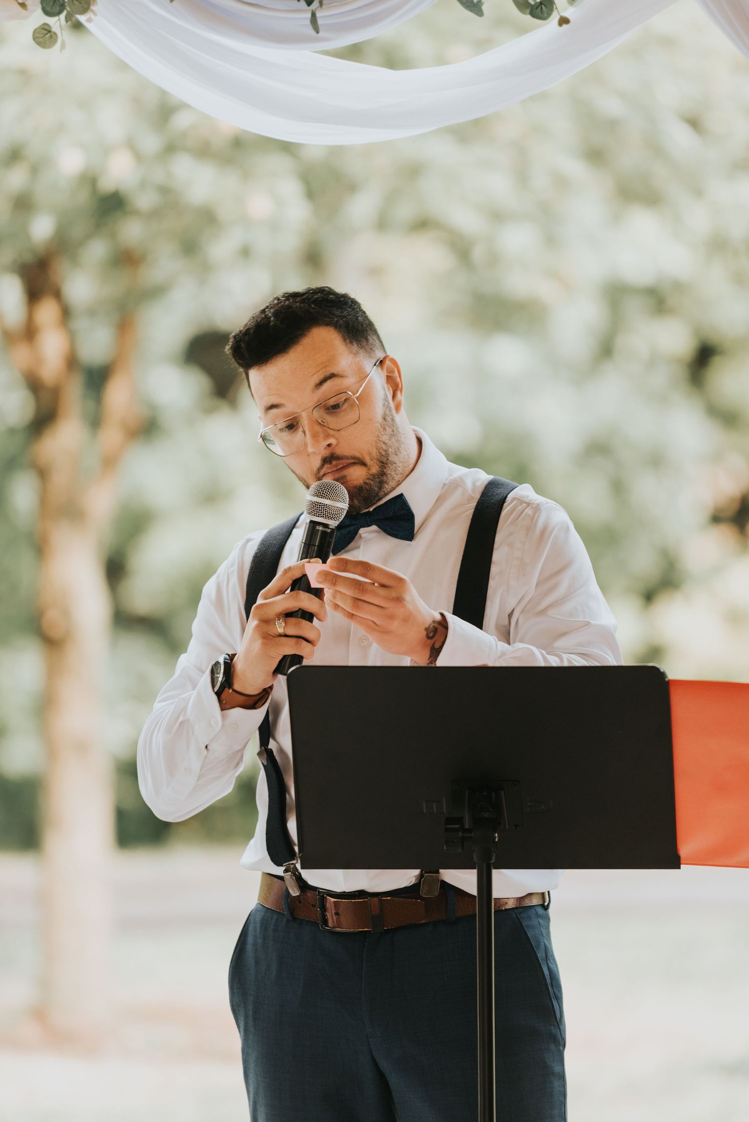 A wedding officiant in suspenders and bow tie reads from notes at an outdoor ceremony.
