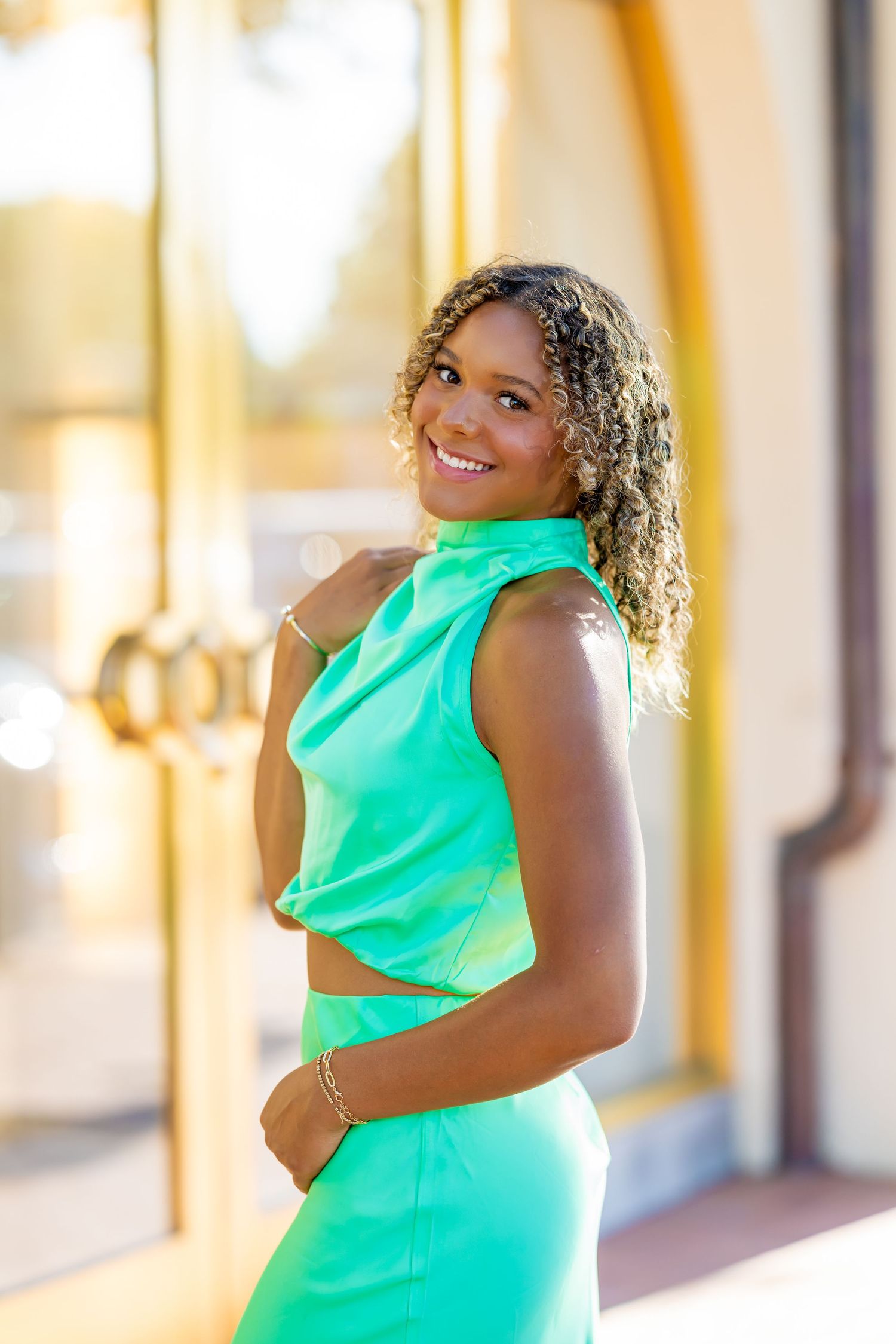 A person in a bright mint green sleeveless dress poses cheerfully against a warm-toned building exterior.