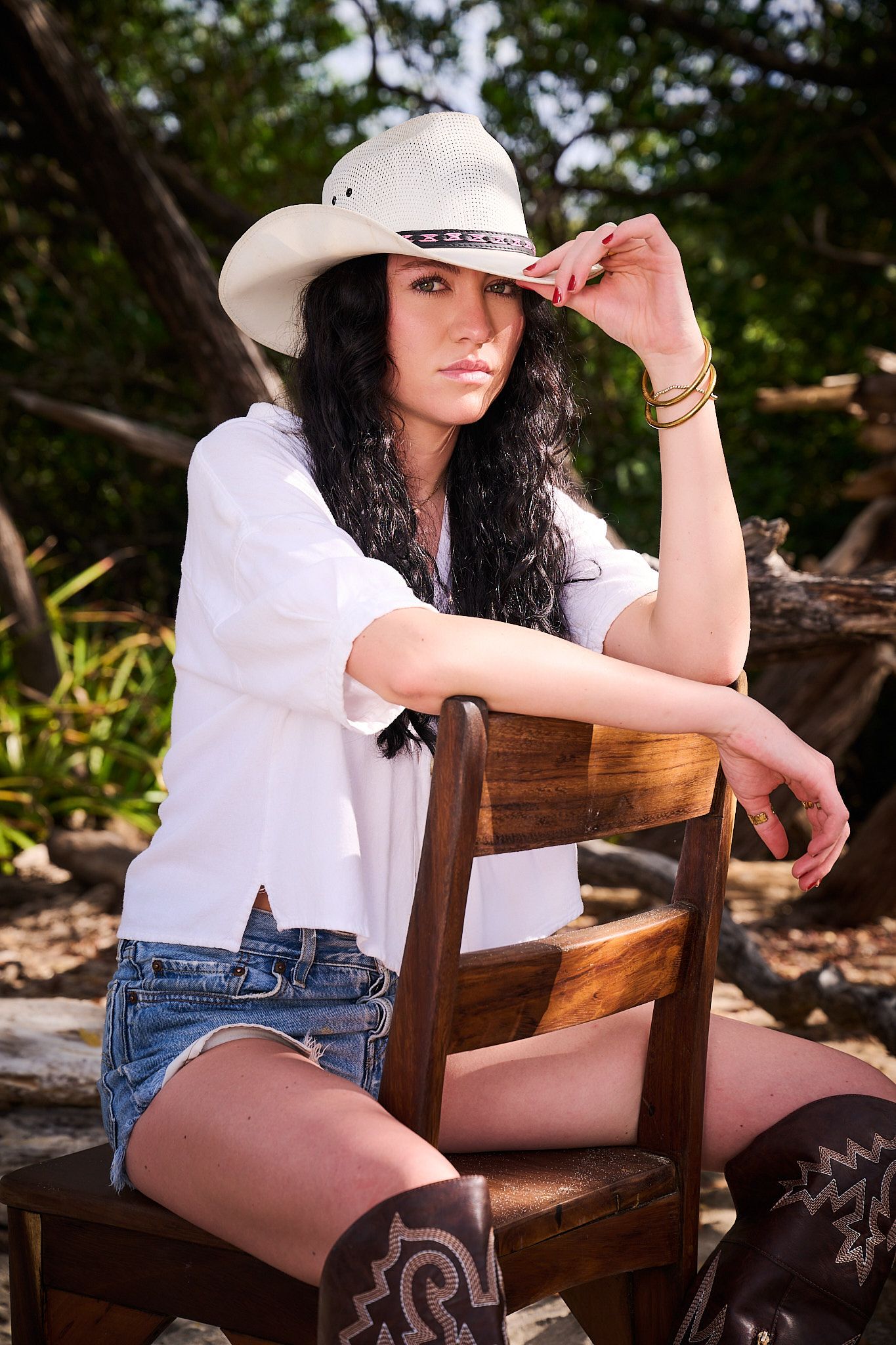 A person in a white shirt, denim shorts and cowboy hat poses on a rustic wooden bench in an outdoor setting.