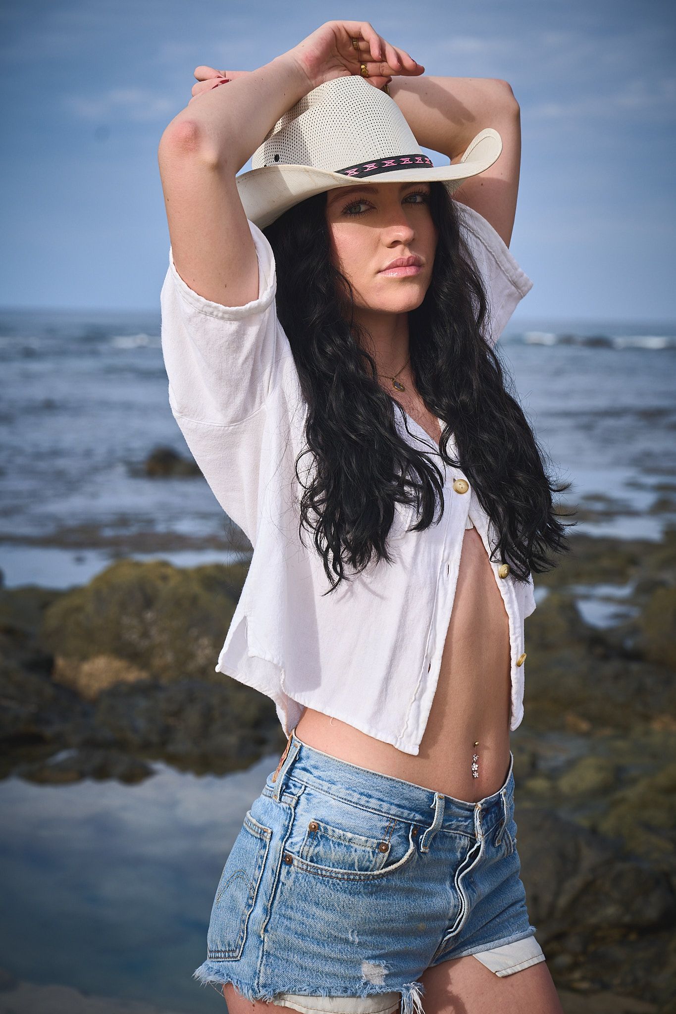 Woman in white shirt and denim shorts poses at rocky beach wearing cowboy hat with ocean in background.