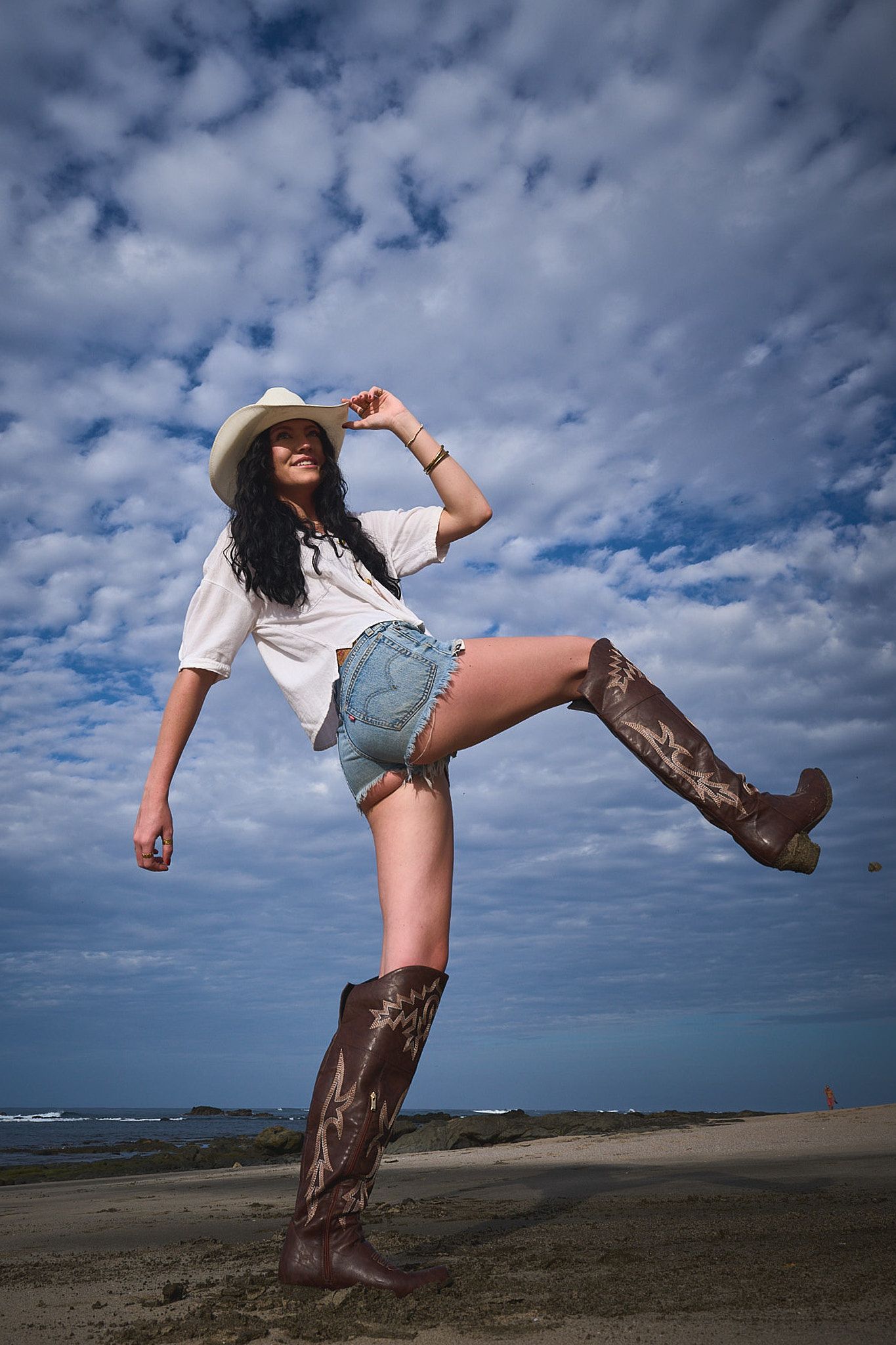 A person in cowboy boots, denim shorts and white shirt poses playfully against a dramatic cloudy sky at the beach.