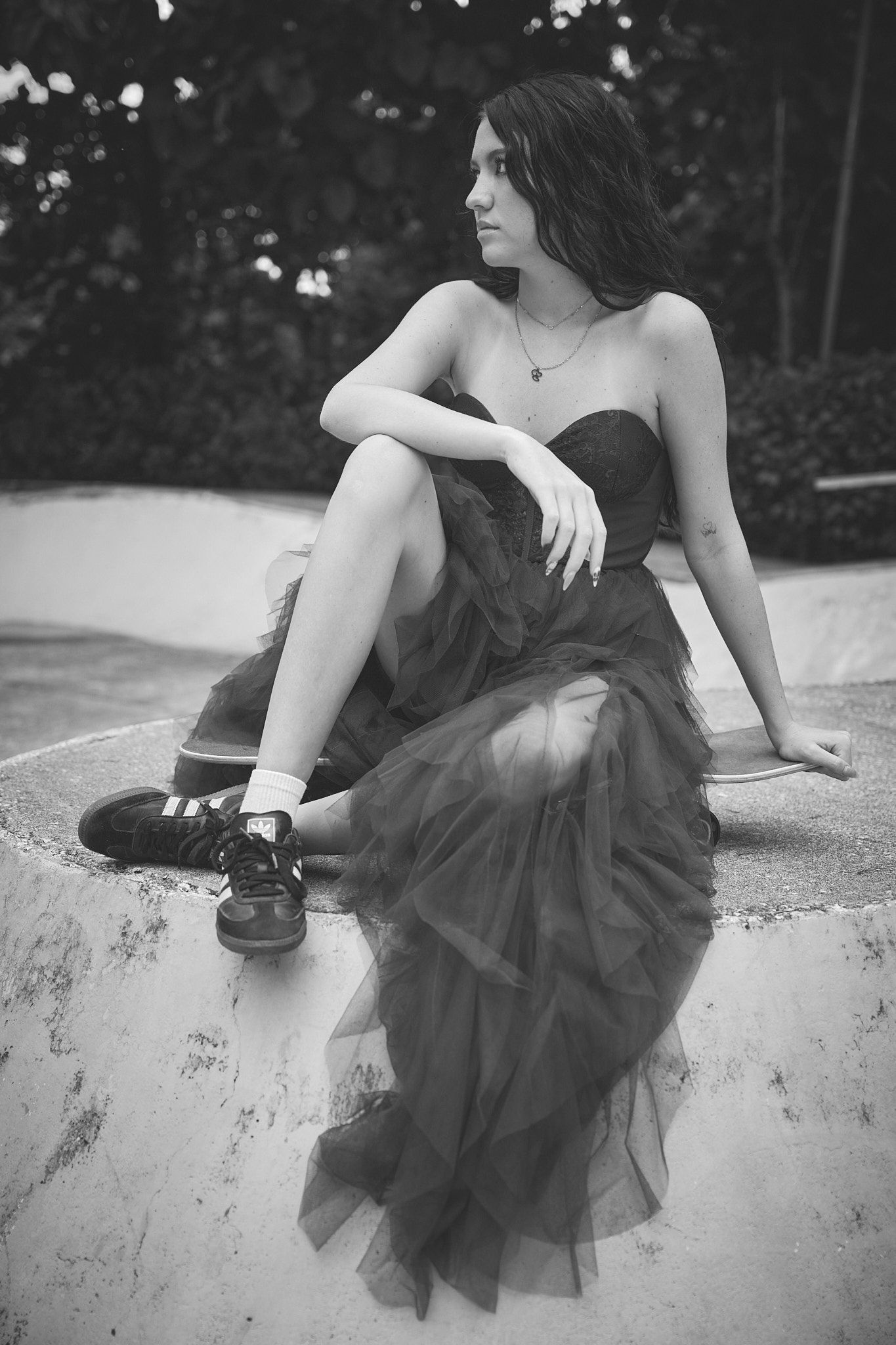 A figure in a dark tulle dress sits gracefully along a concrete wall in a black and white artistic photo series.
