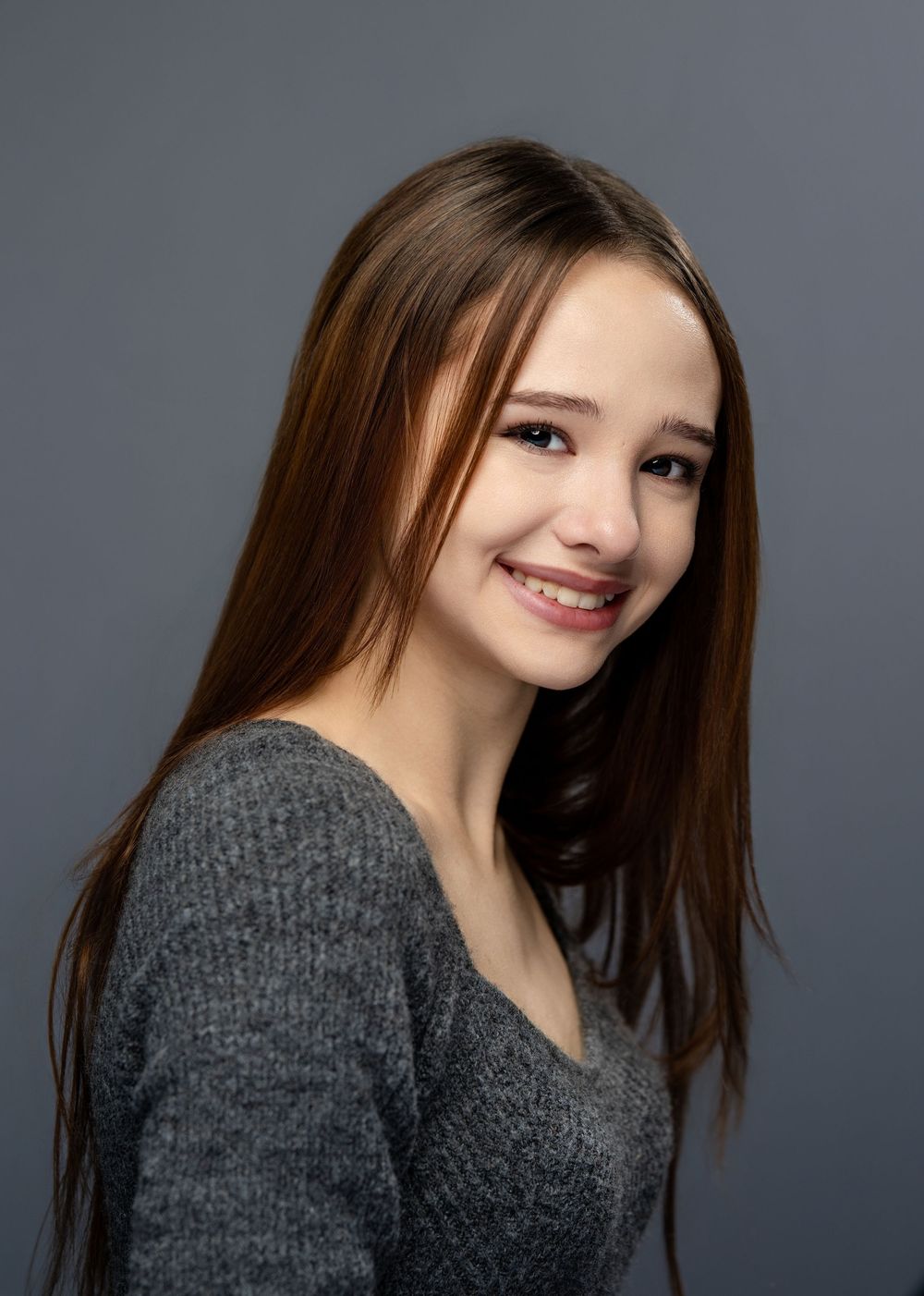 Young girl with long brown hair smiling warmly in a gray sweater against a gray studio background.