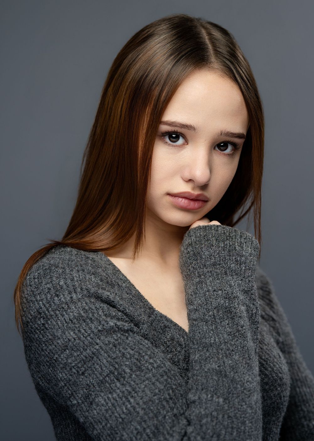 Young woman with long brown hair in a gray sweater poses for a close-up portrait.